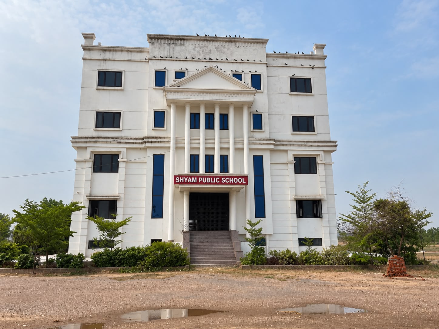 The main entrance of Shyam Public School — a white, pillared portico with the school's name board above the doorway, reached by a broad flight of steps