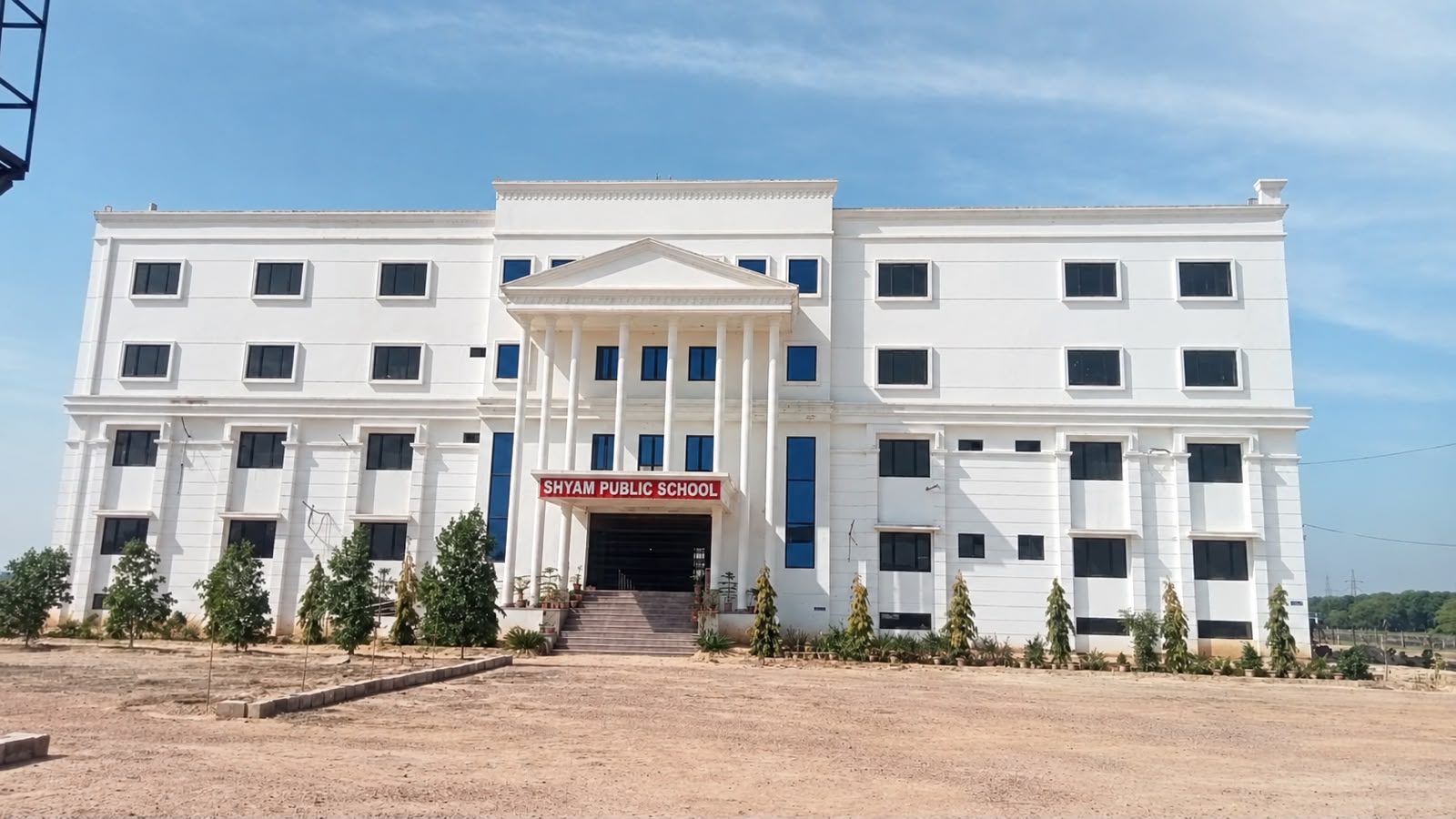 The full front elevation of Shyam Public School: a long three-storey white building with a central pillared portico and a red name board, with young trees planted along the approach